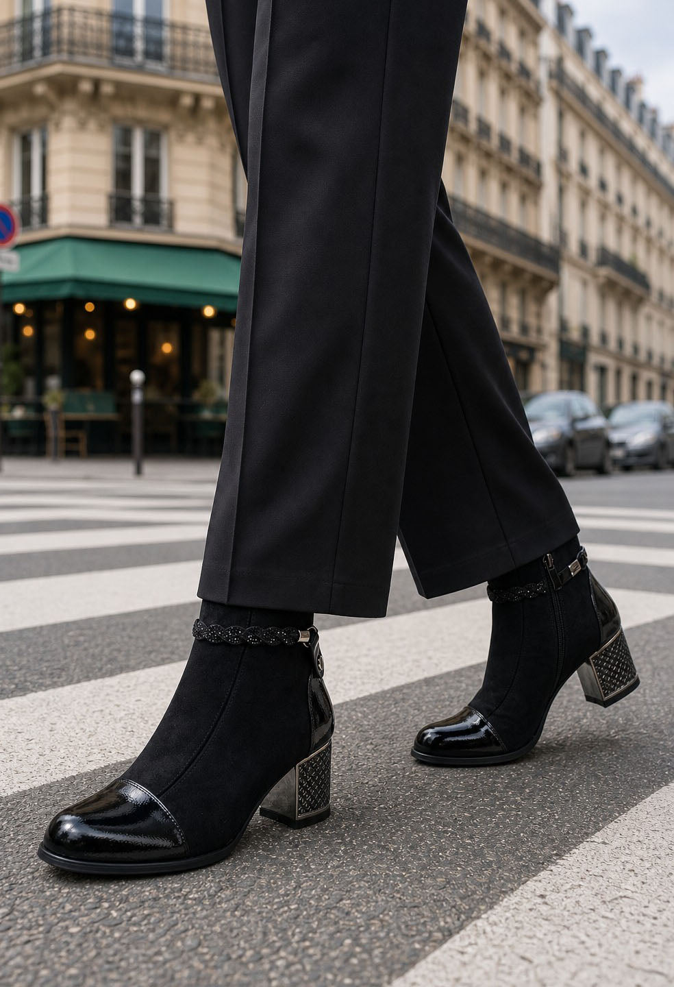 Black ankle boots with a glossy patent leather toe cap and a textured metallic block heel, worn with black trousers on a Parisian street crosswalk.