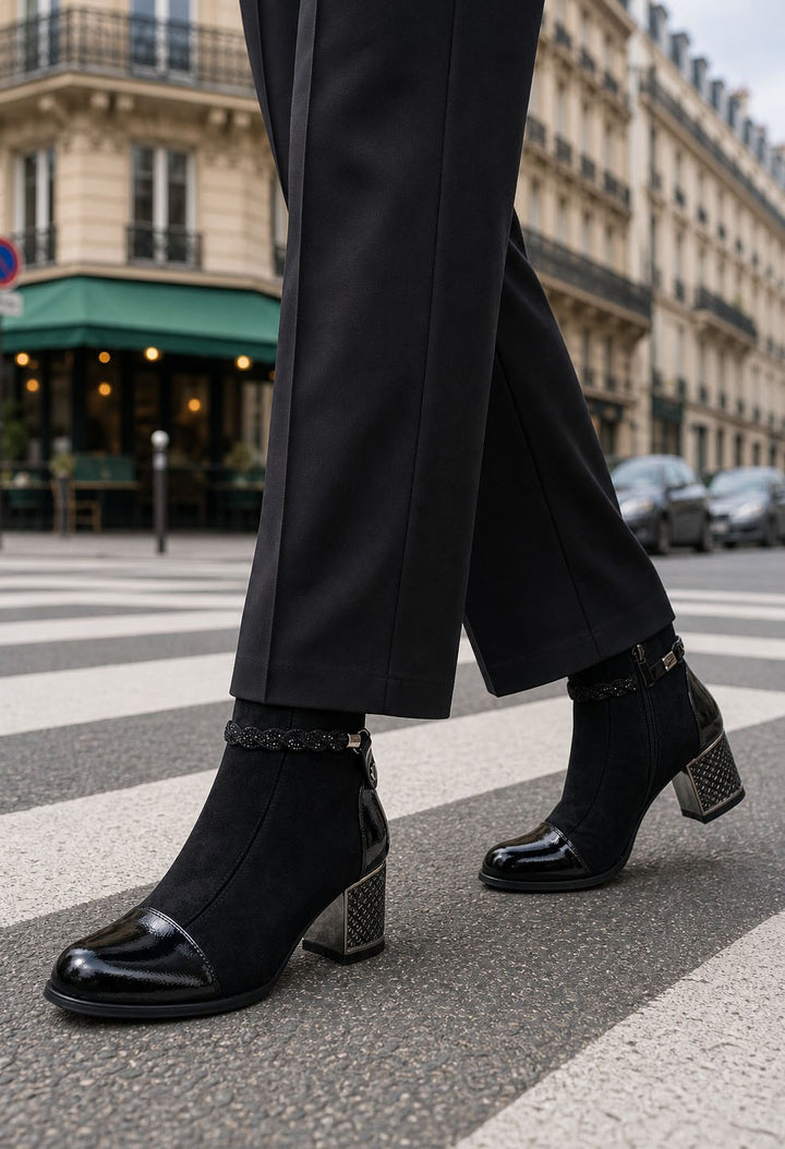 Black ankle boots with a glossy patent leather toe cap and a textured metallic block heel, worn with black trousers on a Parisian street crosswalk.