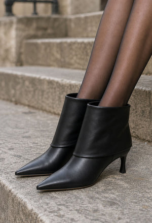 Black leather pointed-toe ankle boots with kitten heels, shown on a model's feet wearing sheer black tights while sitting on stone steps.