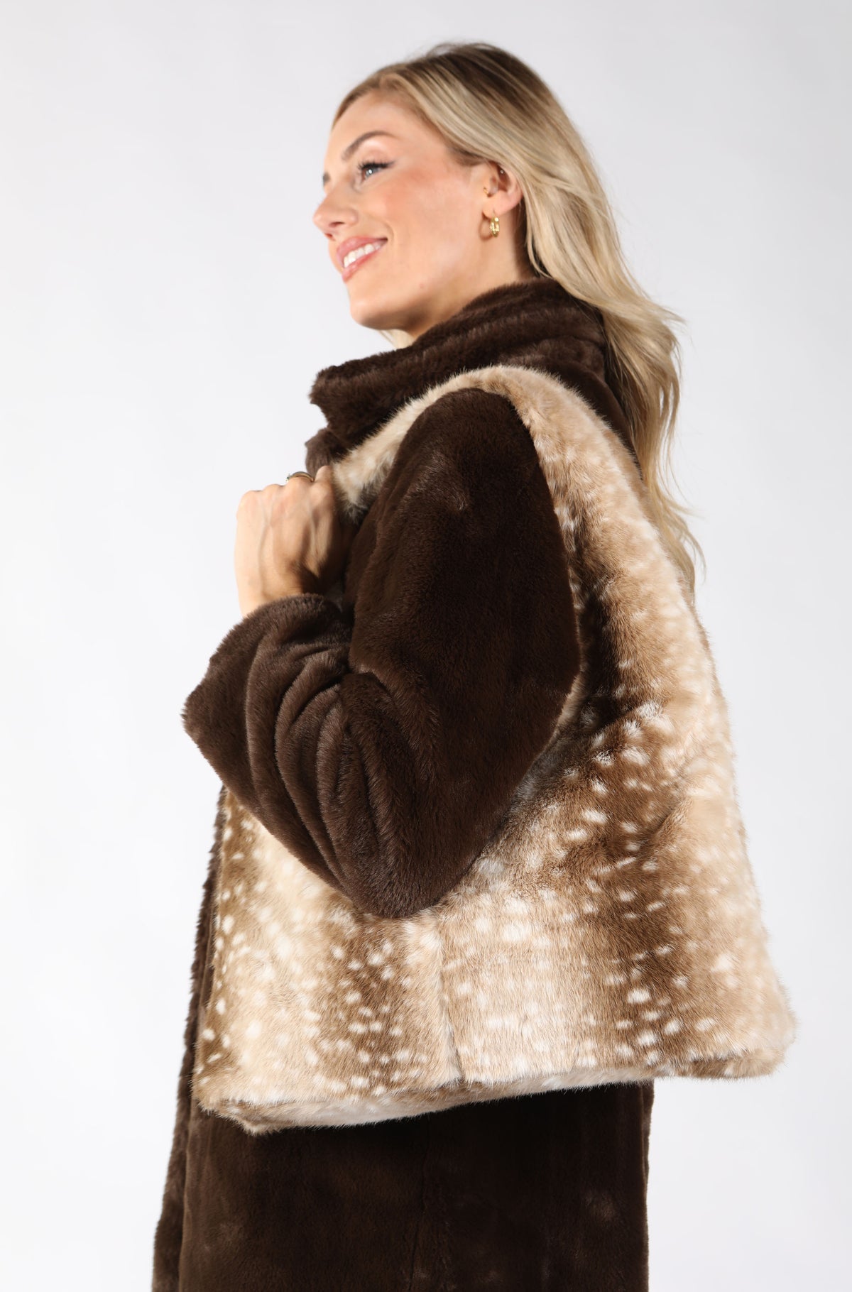 A woman wearing a brown and beige faux fur vest with white spotted accents, paired with a dark brown top, photographed against a plain white studio background.