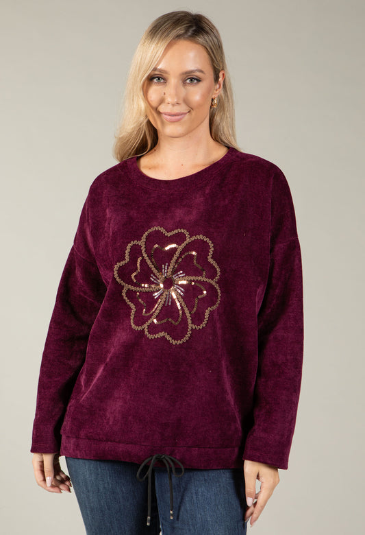 A woman wearing a deep burgundy long-sleeve crewneck sweater with a gold sequined floral design on the front, paired with blue jeans, against a solid light gray background.
