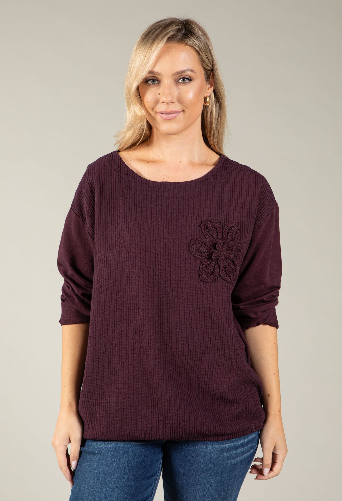 A woman wearing a dark plum ribbed knit top with a tonal embroidered flower on the chest, paired with blue jeans, against a solid light gray background.