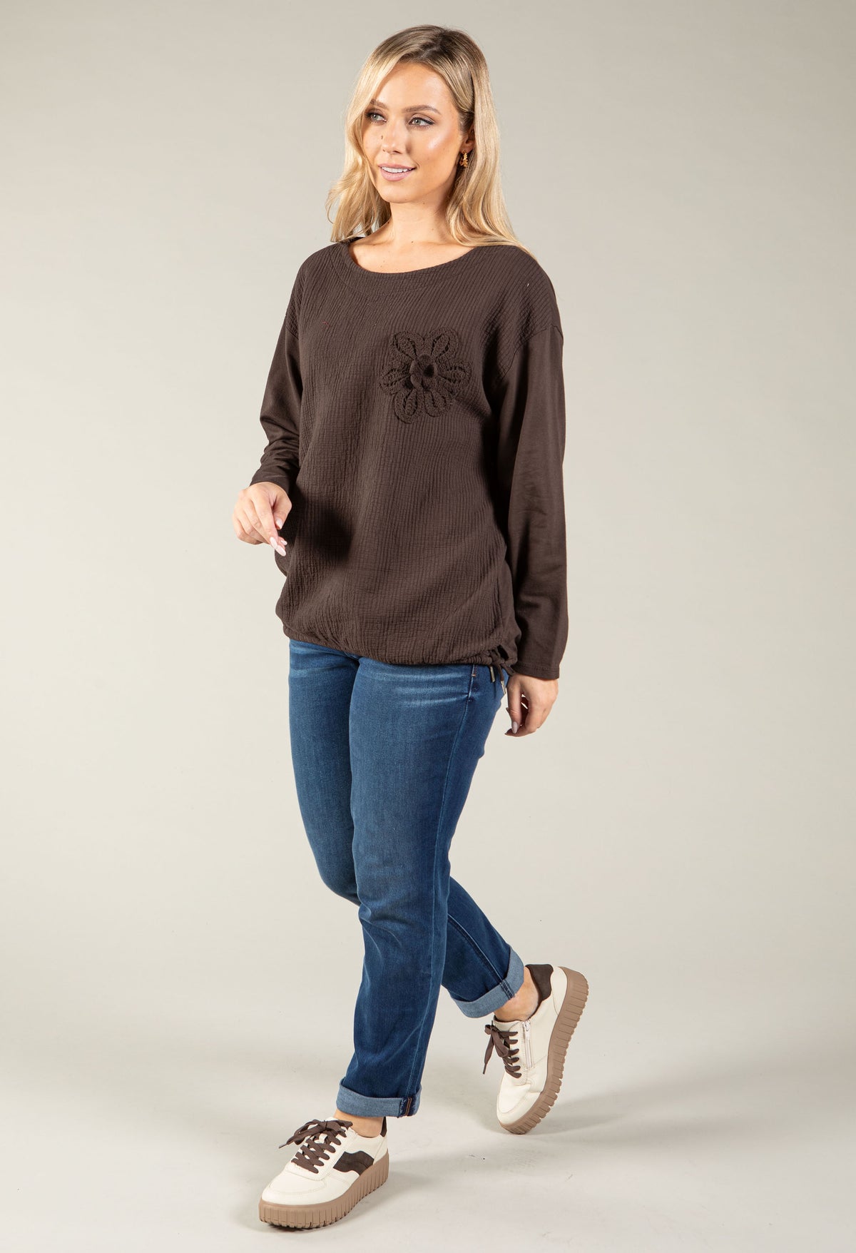 A woman modeling a dark brown long-sleeve knit top with a textured flower detail on the chest, paired with blue jeans and brown-and-white sneakers, against a solid light gray studio background.