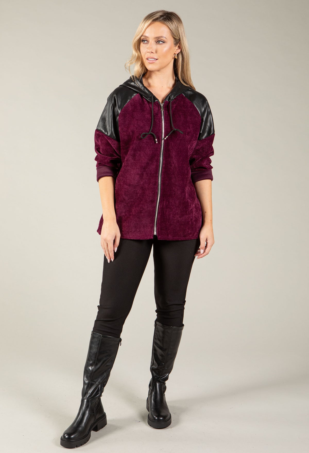 A woman modeling a zip-up hoodie with a deep burgundy suede-like body and black faux-leather sleeves and hood, paired with black pants and tall black boots against a light gray studio background.