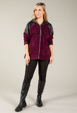 A woman modeling a zip-up hoodie with a deep burgundy suede-like body and black faux-leather sleeves and hood, paired with black pants and tall black boots against a light gray studio background.