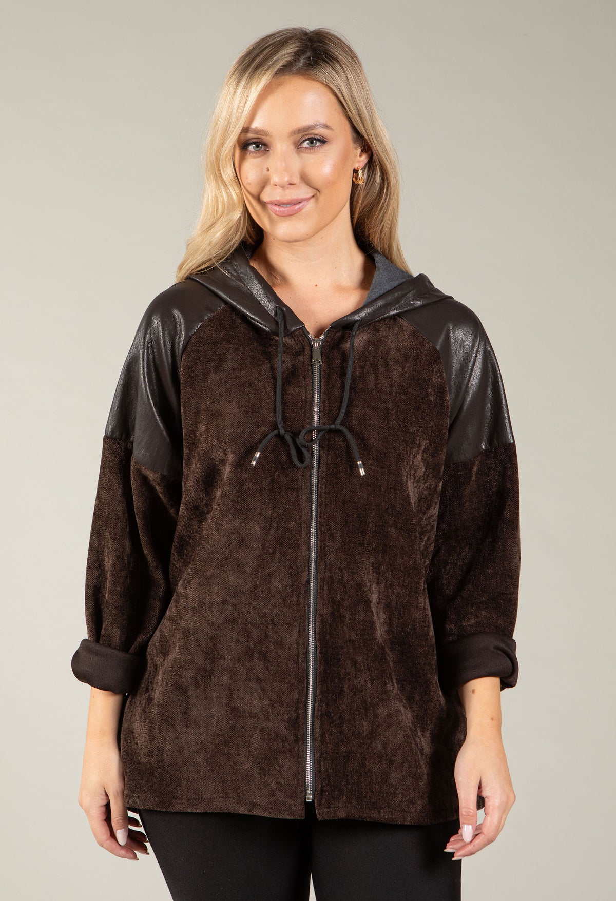 A woman wearing a brown hooded zip-up jacket with dark brown faux-leather sleeves and hood, paired with black pants, against a solid light gray studio background.