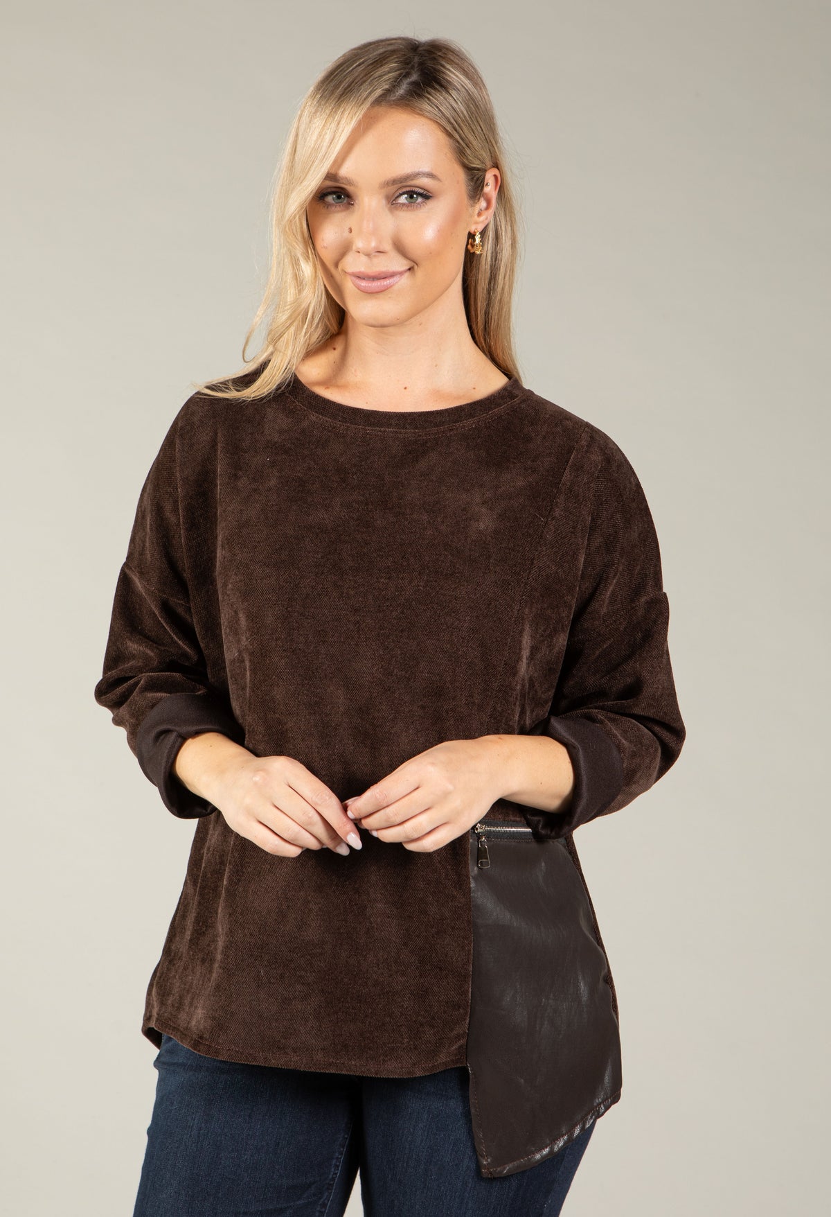A woman modeling a dark brown long-sleeve top with a textured suede-like finish and an asymmetrical dark brown panel on the lower front, paired with dark blue jeans against a solid light gray background.