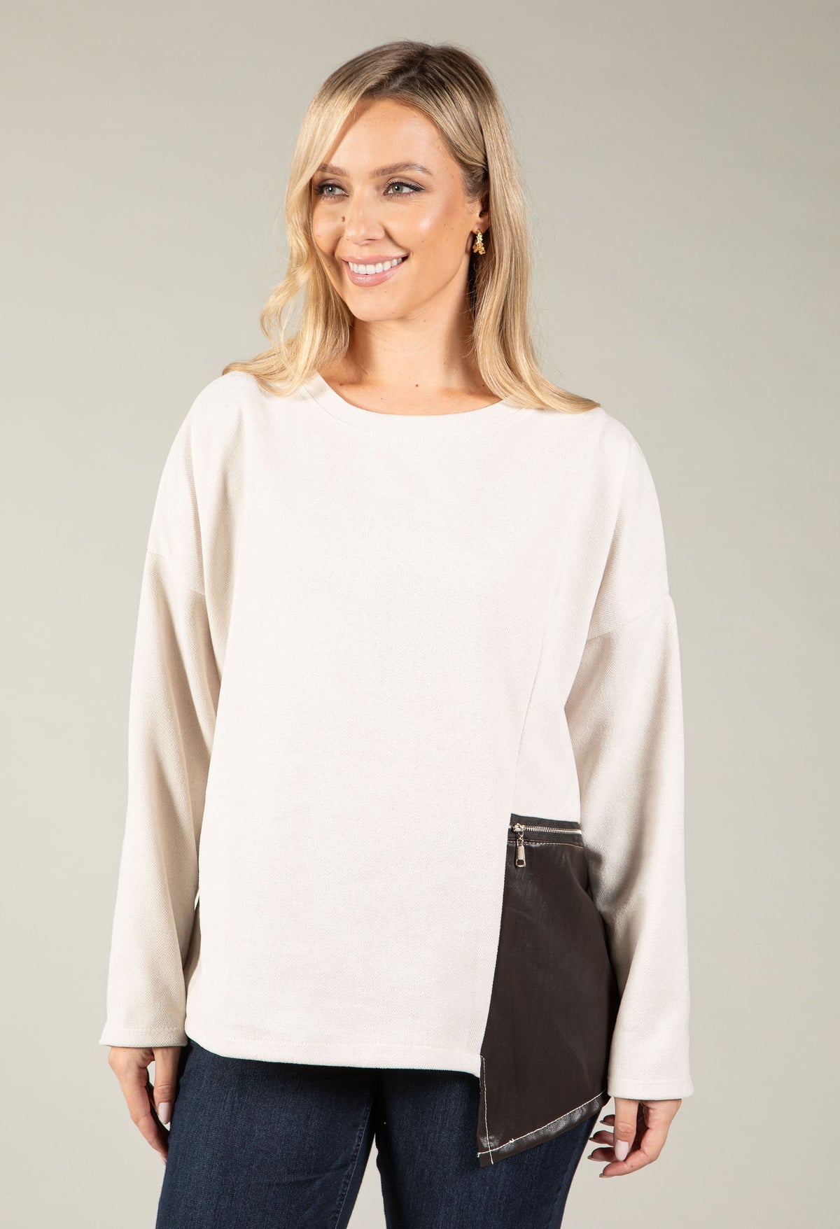 A woman modeling a cream-colored long-sleeve top with an asymmetrical dark brown pocket detail on the lower front, paired with dark blue jeans, against a solid light gray background.