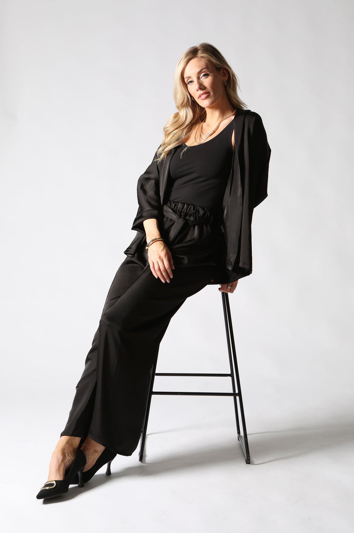 A woman posing in a black three-piece outfit consisting of a scoop-neck top, wide-leg trousers, and a matching open-front jacket draped over her shoulders, seated on a black metal stool against a light gray studio background.