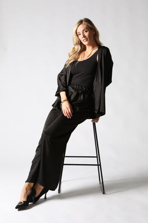 A woman posing in a black three-piece outfit consisting of a scoop-neck top, wide-leg trousers, and a matching open-front jacket draped over her shoulders, seated on a black metal stool against a light gray studio background.