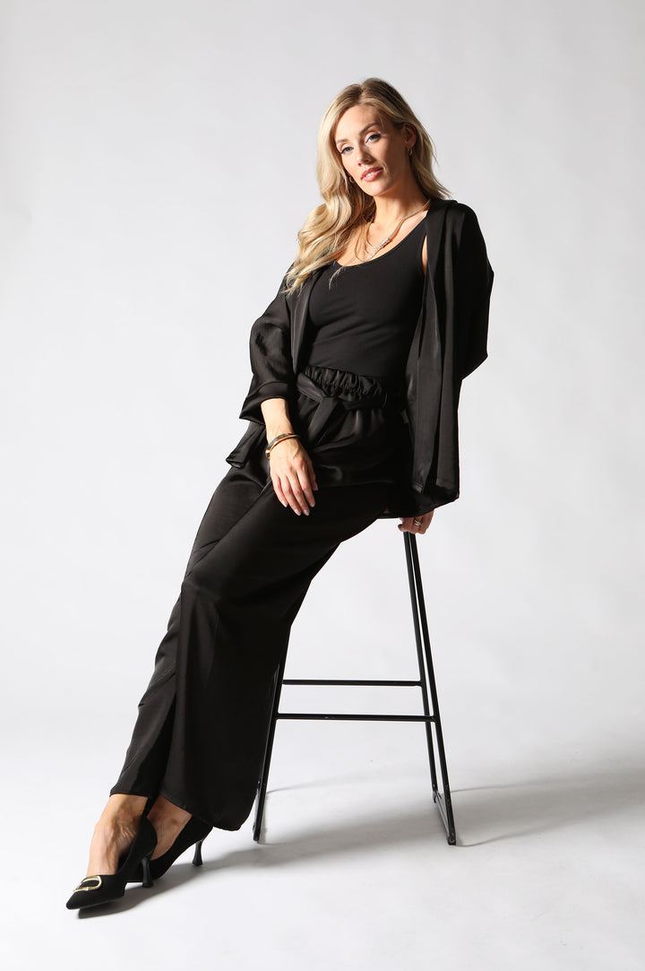 A woman posing in a black three-piece outfit consisting of a scoop-neck top, wide-leg trousers, and a matching open-front jacket draped over her shoulders, seated on a black metal stool against a light gray studio background.