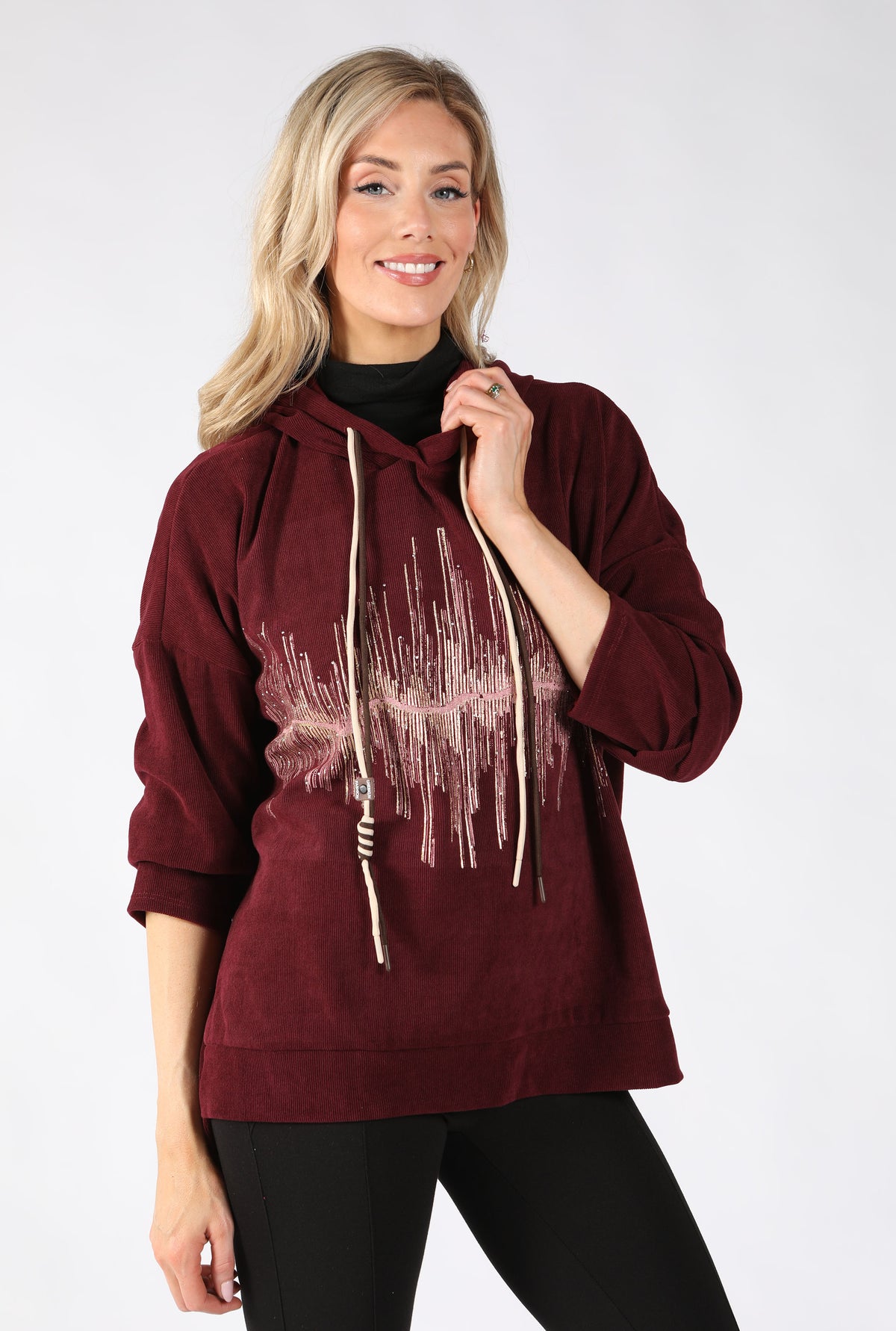 A woman modeling a burgundy hooded sweatshirt with a distressed graphic print on the front, featuring cream-colored drawstrings, paired with black pants against a light gray studio background.