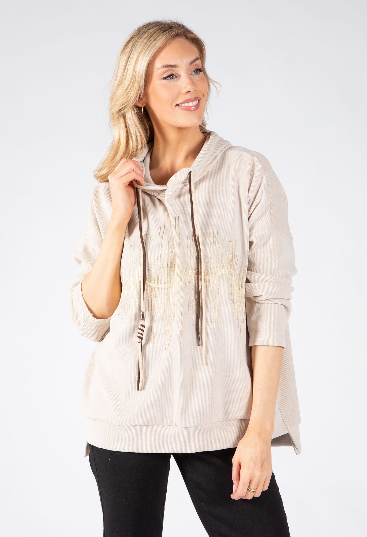 A woman modeling a light beige hooded sweatshirt with fringe detailing on the front, paired with black pants, against a plain white studio background.
