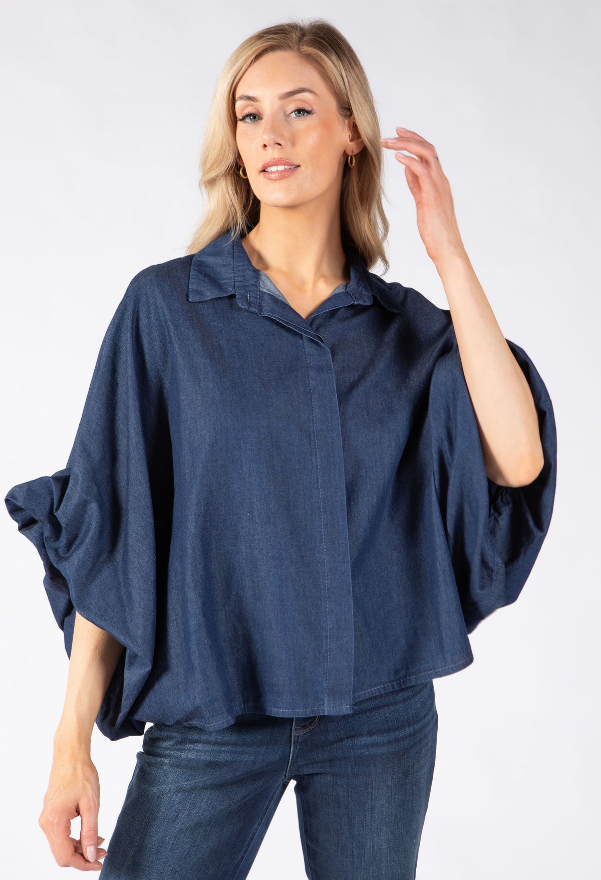 A woman modeling a dark blue, loose-fitting button-up blouse with wide and draped sleeves.