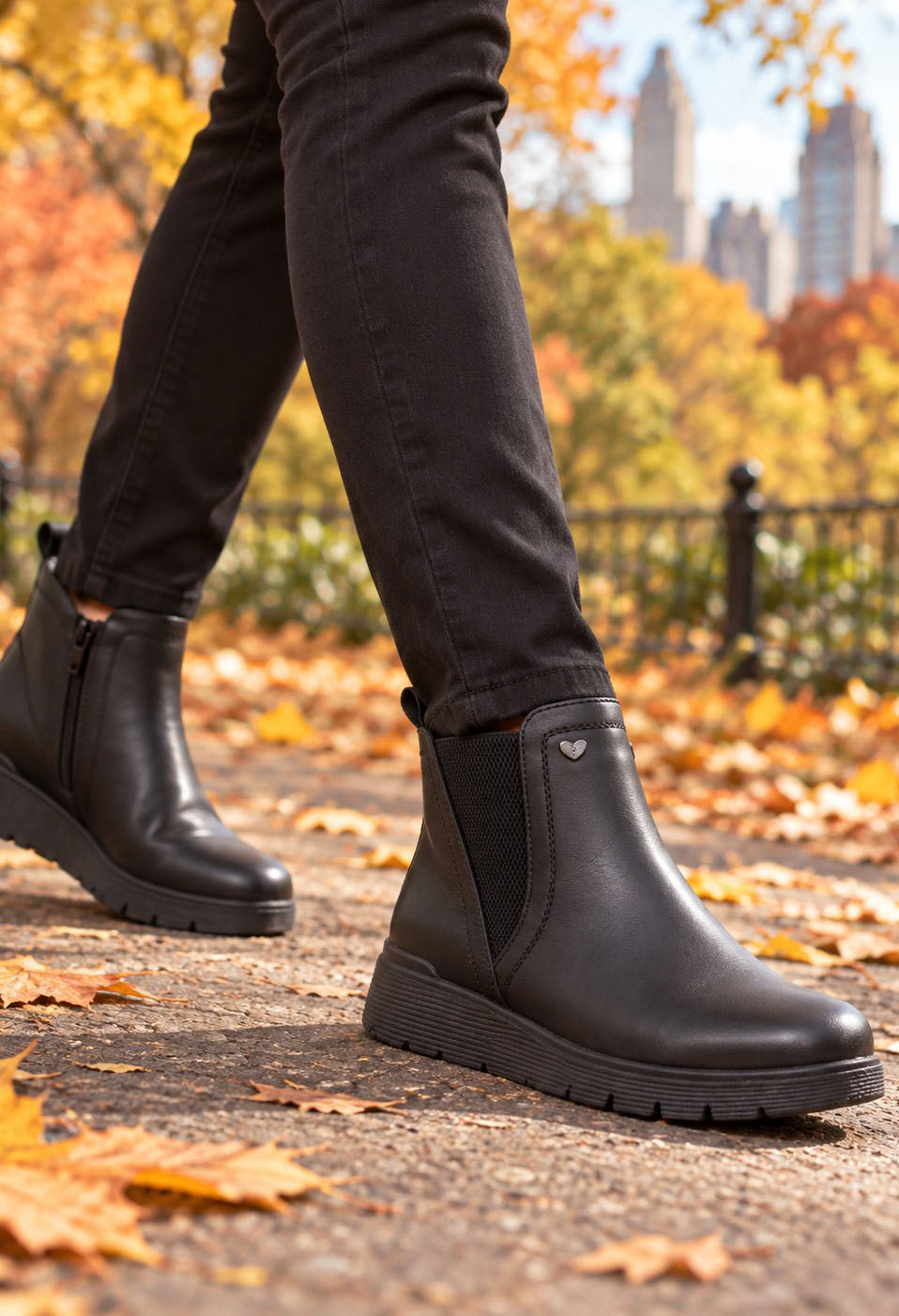 Black leather Chelsea-style ankle boots worn with black jeans, shown in an outdoor autumn park setting with fallen leaves and city buildings in the background.