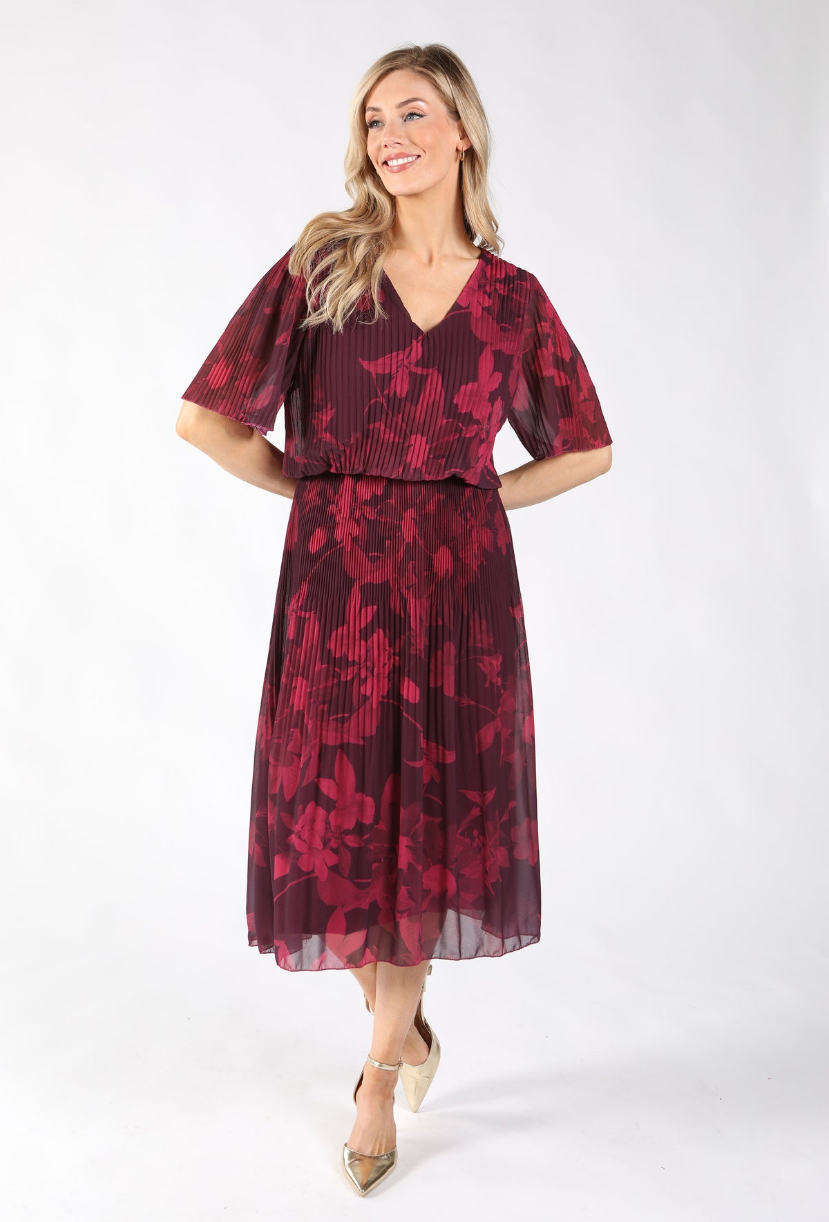 A woman modeling a pleated midi dress with a deep V-neck and flutter sleeves. The dress features a dark burgundy base with a red floral pattern, paired with subtle vertical pleating.