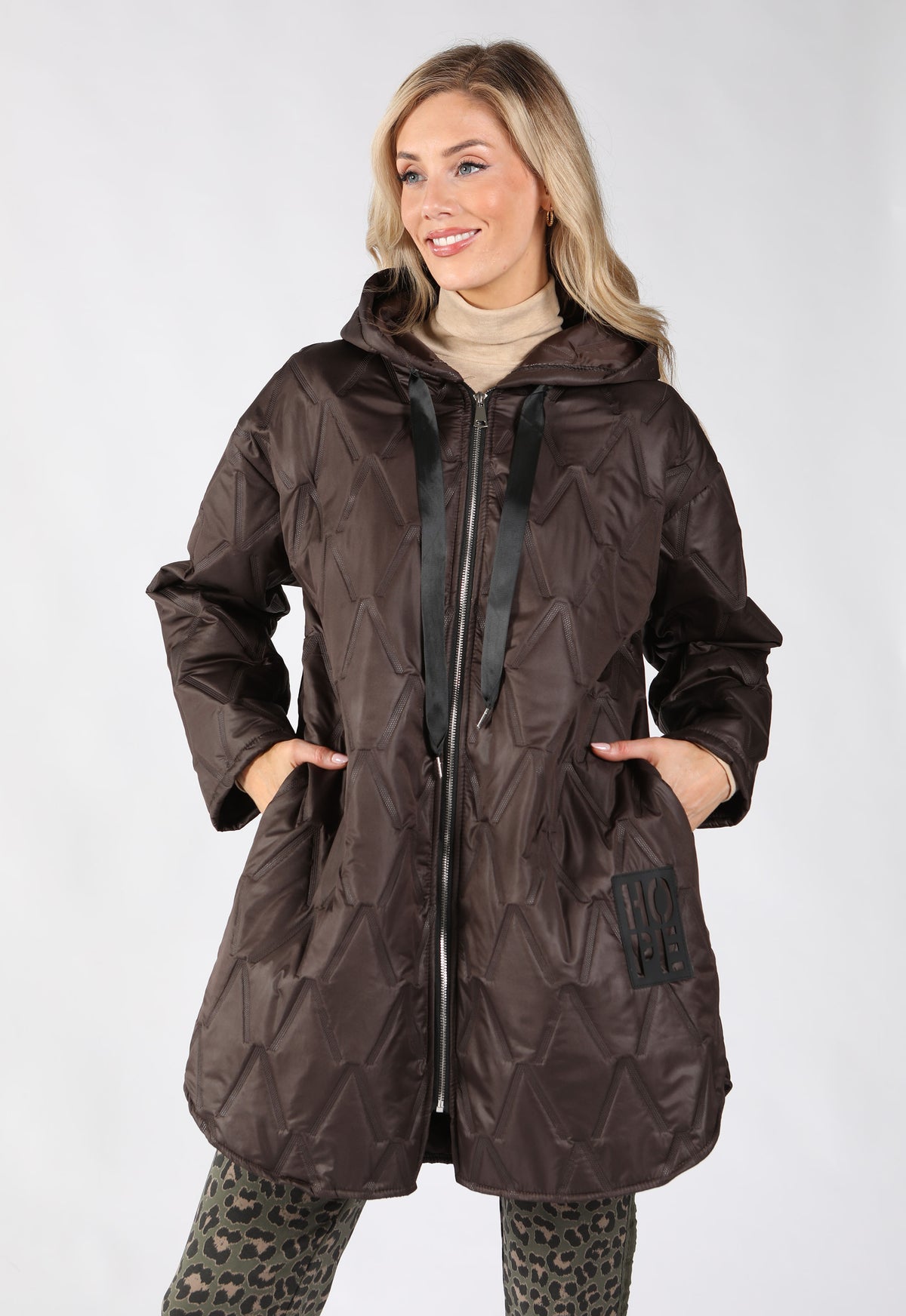 A woman modeling a dark brown quilted hooded jacket with a front zipper, black drawstrings, and a kangaroo pocket.