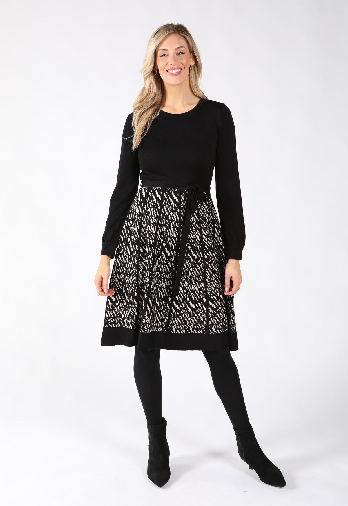 Model wearing knitted dress with long sleeves and a standout zig-zag patterned skirt in ivory and black. Cinched with a belt tie.