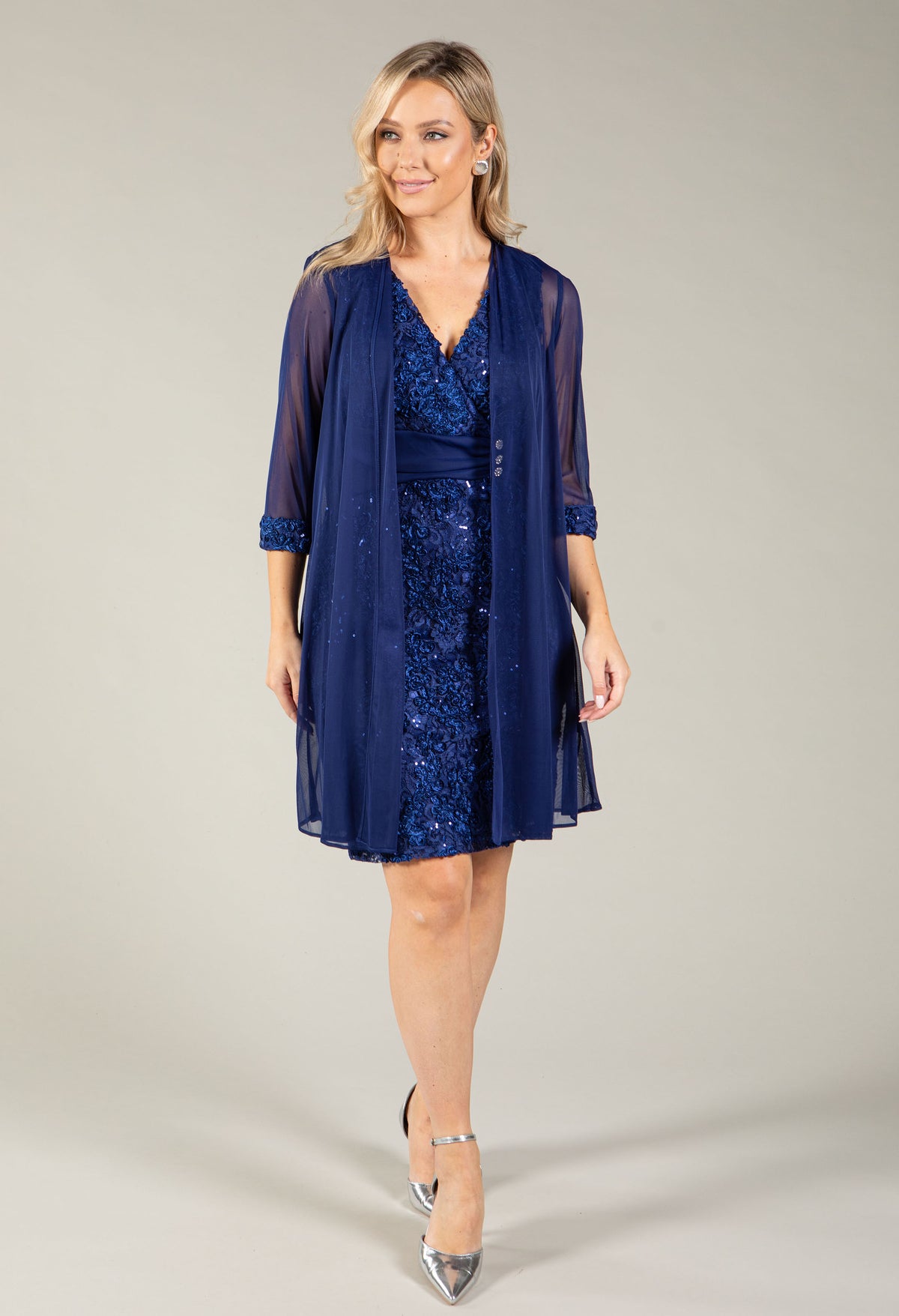A woman modeling a navy blue three-piece formalwear set consisting of a V-neck sequined dress, a matching sheer embroidered jacket, and silver pointed-toe heels, against a solid light gray studio background.