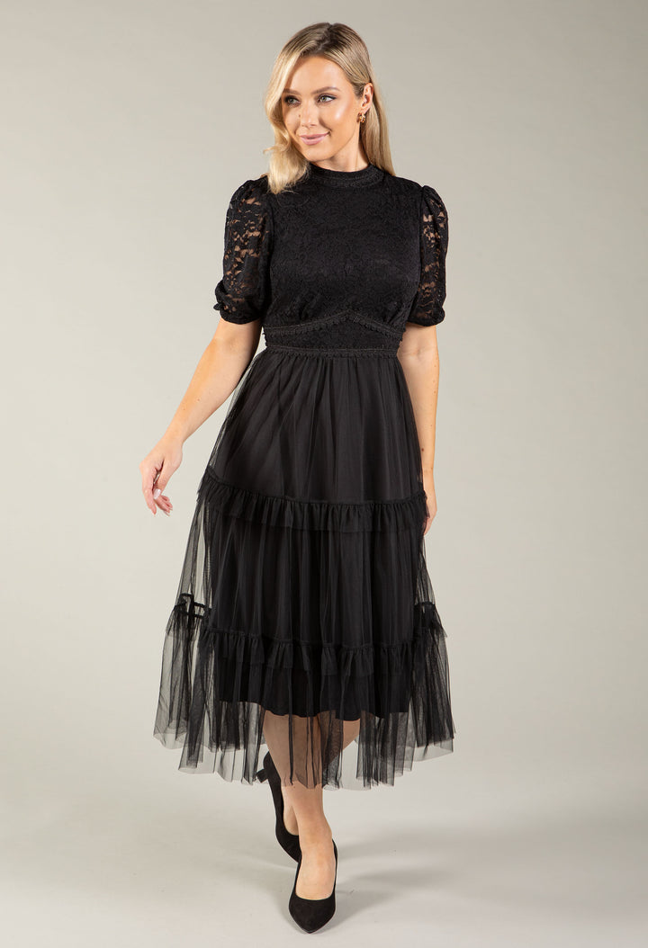 A woman modeling a black midi dress with a lace bodice, short puff sleeves, and a tiered tulle skirt, photographed against a plain light gray studio background.