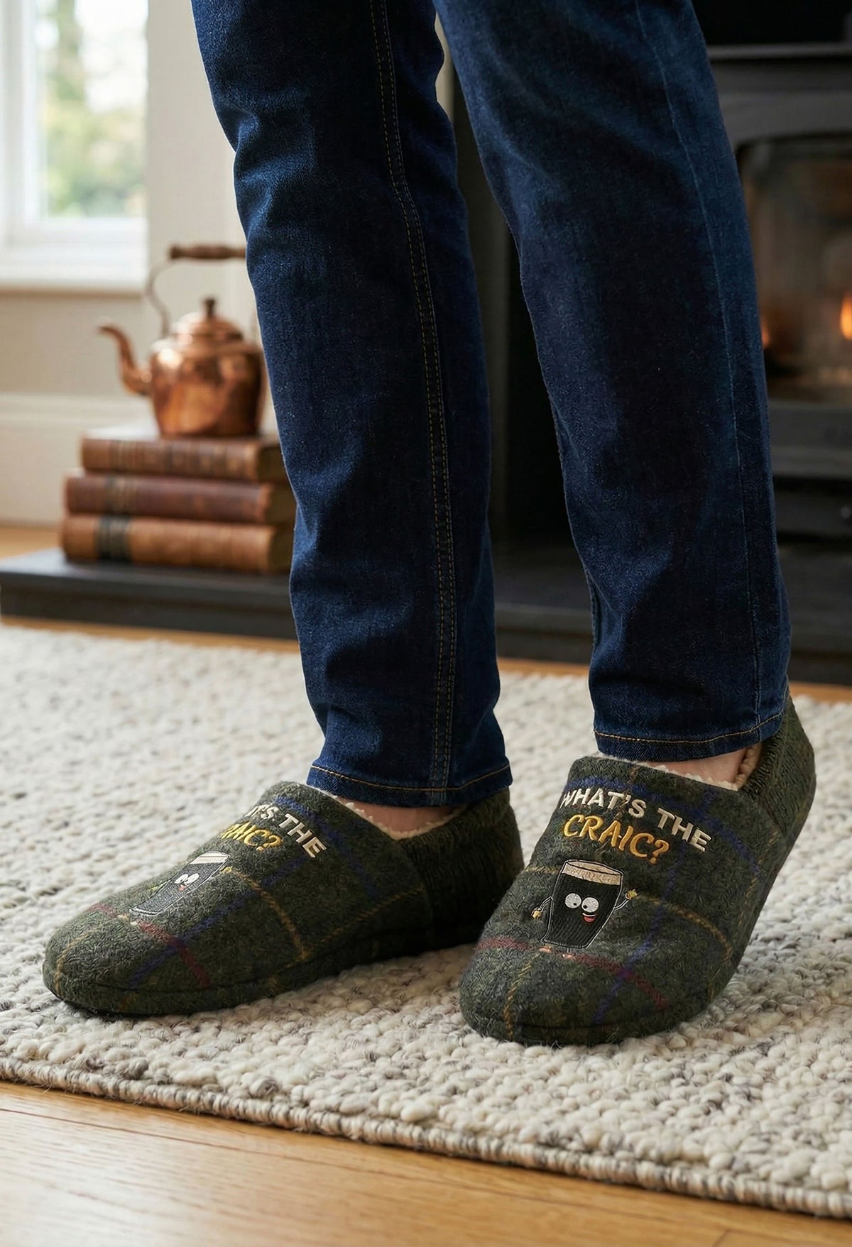 A person wearing dark blue jeans and dark green plaid slip-on slippers with embroidered text and cartoon faces, standing on a light rug in a cozy indoor setting.