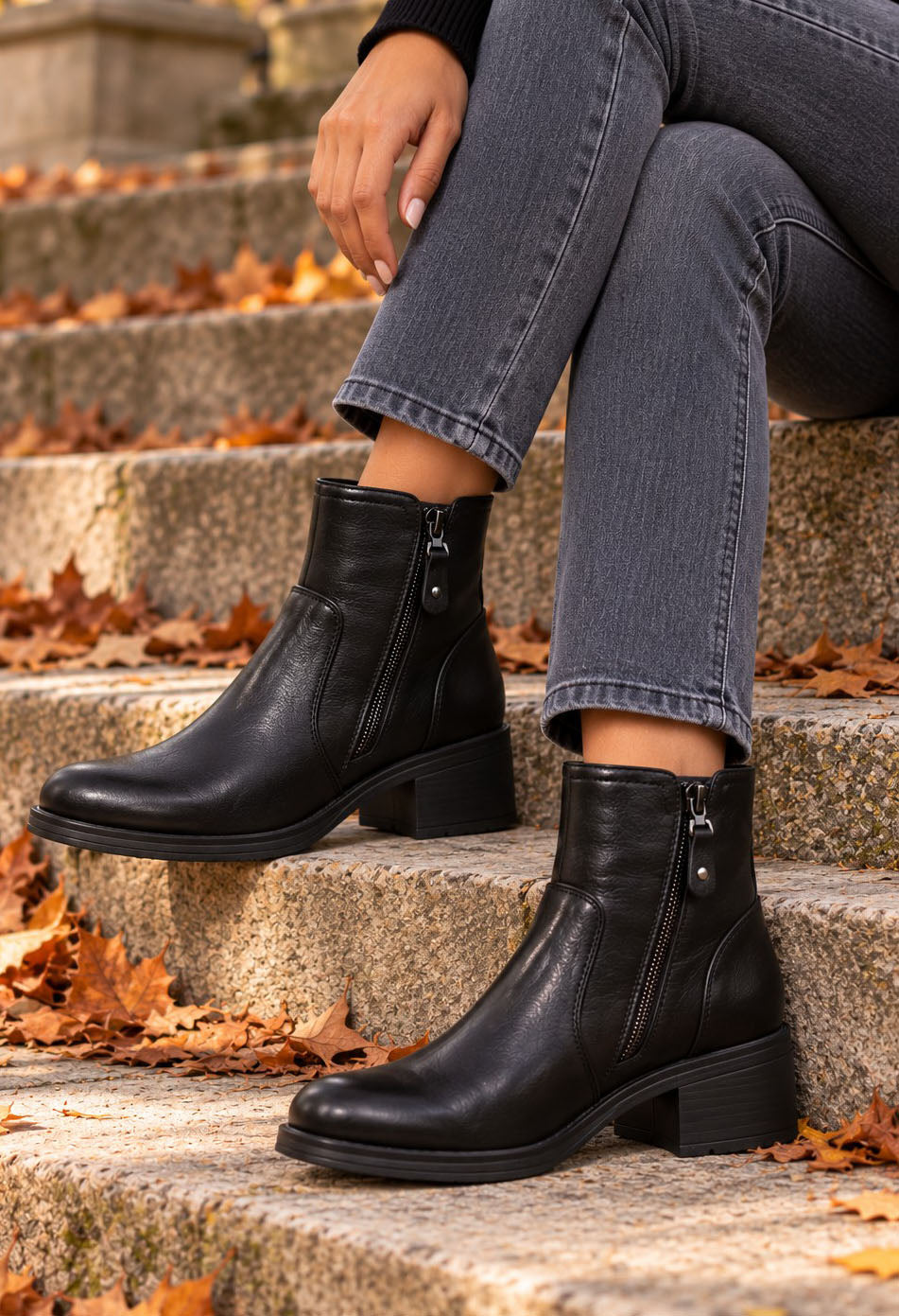 A person sitting on outdoor stone steps wearing black ankle boots with side zippers, gray jeans, and a black top. Fallen autumn leaves are scattered on the steps.