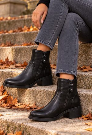 A person sitting on outdoor stone steps wearing black ankle boots with side zippers, gray jeans, and a black top. Fallen autumn leaves are scattered on the steps.