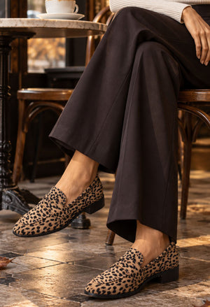 A person seated at a table wearing leopard-print slip-on loafers with dark wide-leg trousers in a cafe setting.