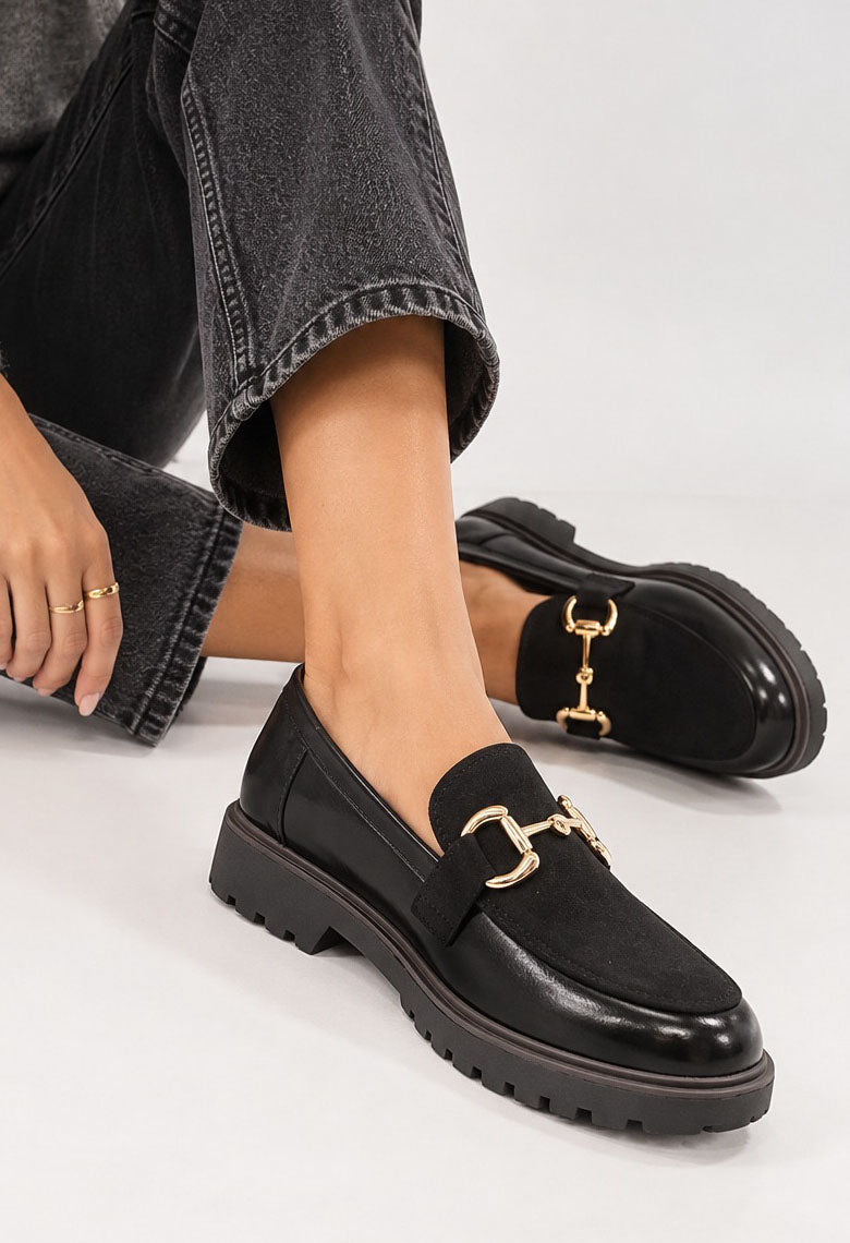 A person wearing black leather loafers with a gold horsebit detail and chunky lug soles, paired with dark gray jeans, on a light gray studio background.