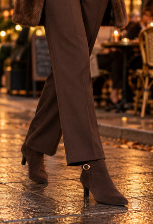 A close-up lifestyle shot of a person's legs walking on a wet cobblestone street at night, wearing brown wide-leg trousers and matching brown suede ankle boots with a small gold buckle detail.