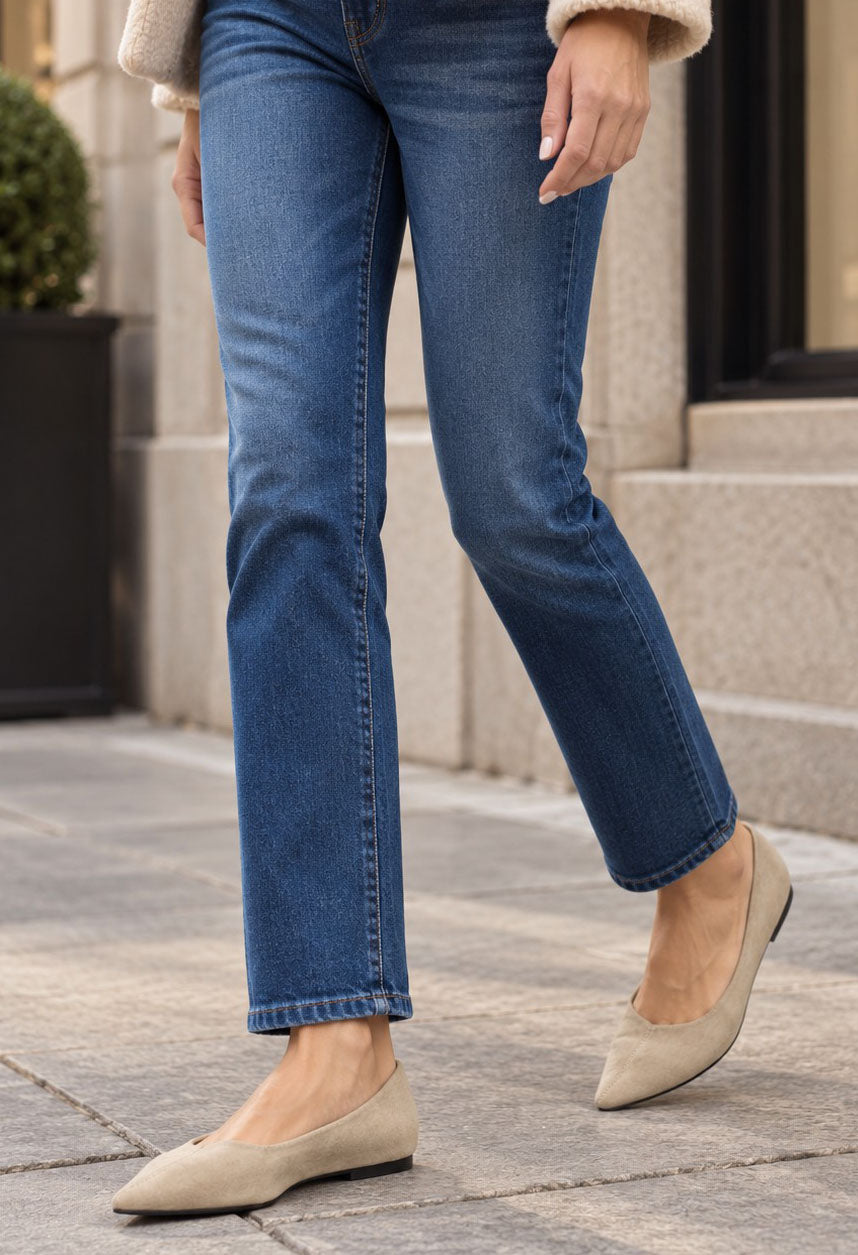 A person wearing medium-wash blue straight-leg jeans and beige pointed flats, standing on a stone sidewalk in front of a building.