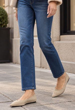 A person wearing medium-wash blue straight-leg jeans and beige pointed flats, standing on a stone sidewalk in front of a building.