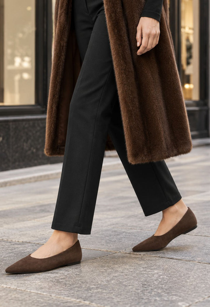 A person walking outdoors wearing a long brown faux-fur coat, black pants, and brown pointed-toe flats on a stone sidewalk.