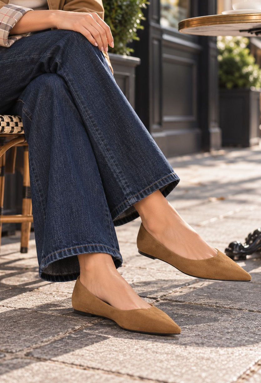 A person seated outdoors wearing pointed-toe flat shoes in a warm camel/tan suede-like material, paired with dark blue wide-leg jeans. The setting appears to be a cafe or street-side seating area with stone pavement and blurred furniture in the background.