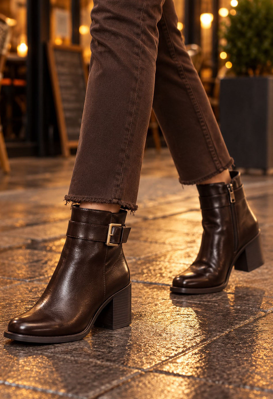 Dark brown leather ankle boots with a side buckle strap and block heel, worn with brown jeans on a wet stone pavement at night.