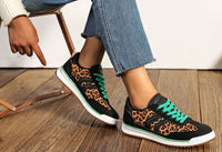 Person wearing black sneakers with orange patterns and green laces on a wooden floor.