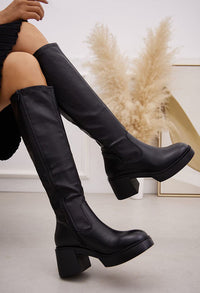 Black knee-high boots worn by a person sitting on the floor with decorative plants in the background.