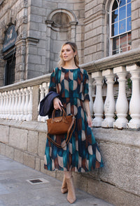Woman wearing patterned dress holding a navy jacket and camel bag