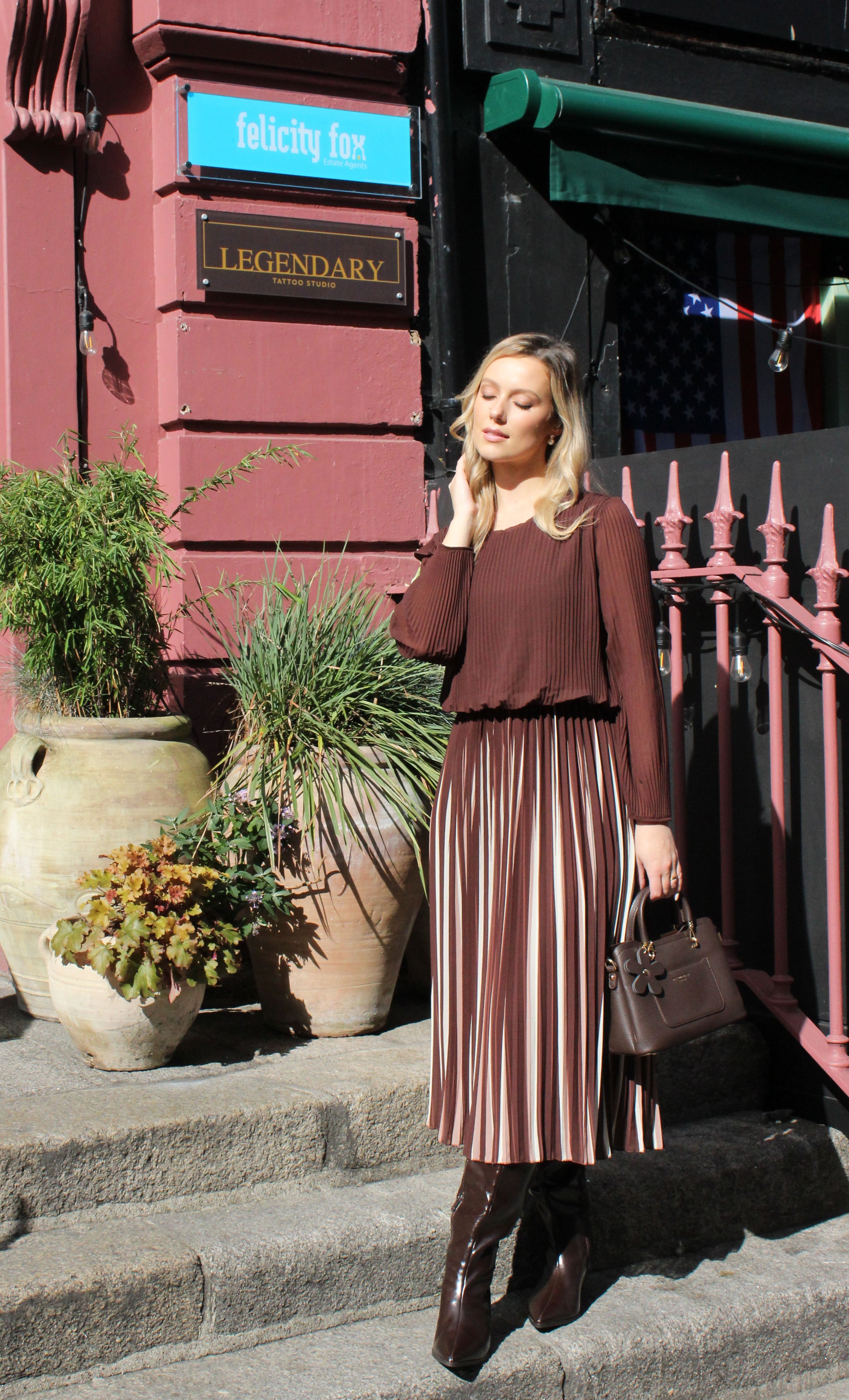 Woman in a brown top and pleated skirt standing on steps with plants and a building in the background