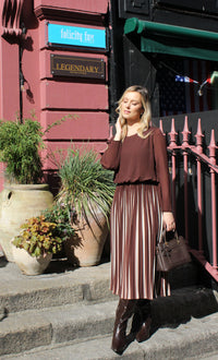 Woman in a brown top and pleated skirt standing on steps with plants and a building in the background