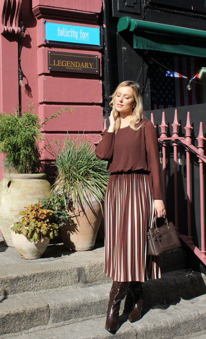 Woman in a brown top and pleated skirt standing on steps with plants and a building in the background