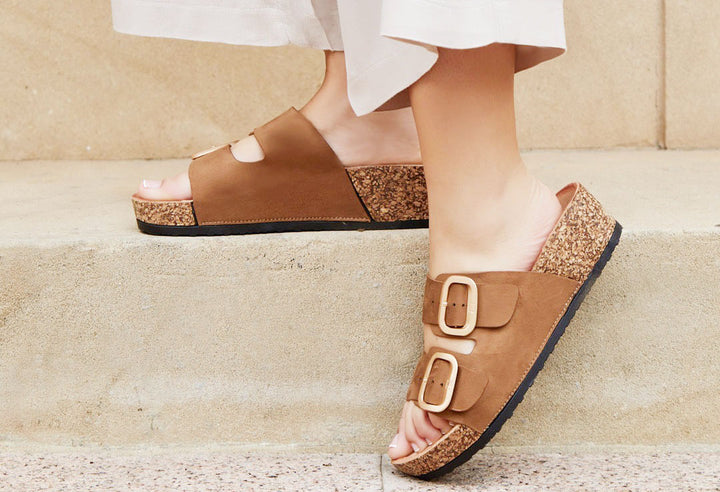 A pair of beige buckle sandals with double straps and a 5cm wedge heel, worn by a person sitting with white pants visible.