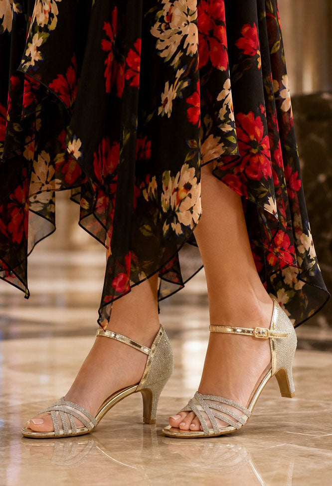 A close-up of a person's feet wearing metallic gold strappy heeled sandals, paired with a black floral dress, standing on a polished marble floor.