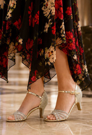 A close-up of a person's feet wearing metallic gold strappy heeled sandals, paired with a black floral dress, standing on a polished marble floor.