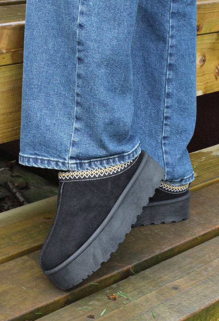 A person wearing blue jeans and black platform slip-on boots with embroidered trim sitting on a wooden bench.