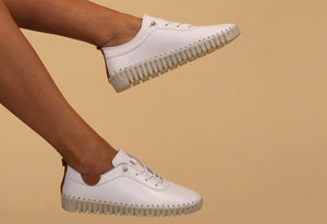 A pair of white leather Plimsoll shoes with a lace-up closure, displayed on a person standing against a white background.