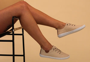 A person seated with their feet hanging off the chair, wearing beige leather plimsolls with lace-up closure.