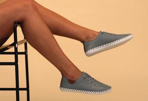 A person seated with their feet hanging off the chair, wearing blue leather plimsolls with lace-up closure.