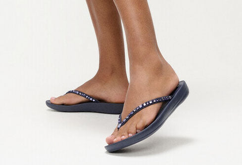 A pair of navy Fit Flop crystal-embellished toe-posts sandals on a model's feet against a white background.