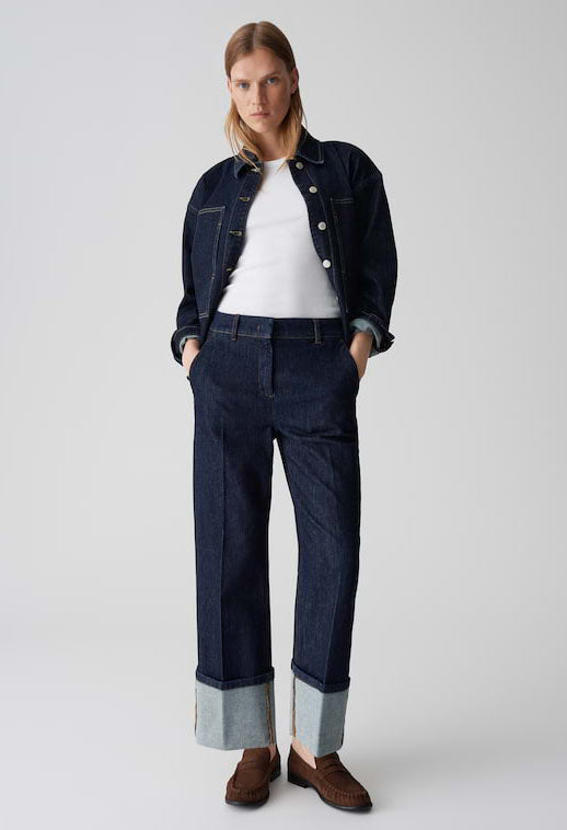 Woman wearing a denim jacket, white shirt, and dark jeans on a plain background